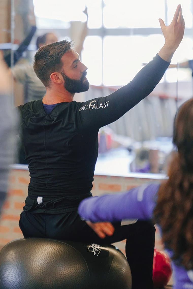 A PBT tutor seated on a stability ball, one arm raised, leading a studio of dance teachers through a conditioning class